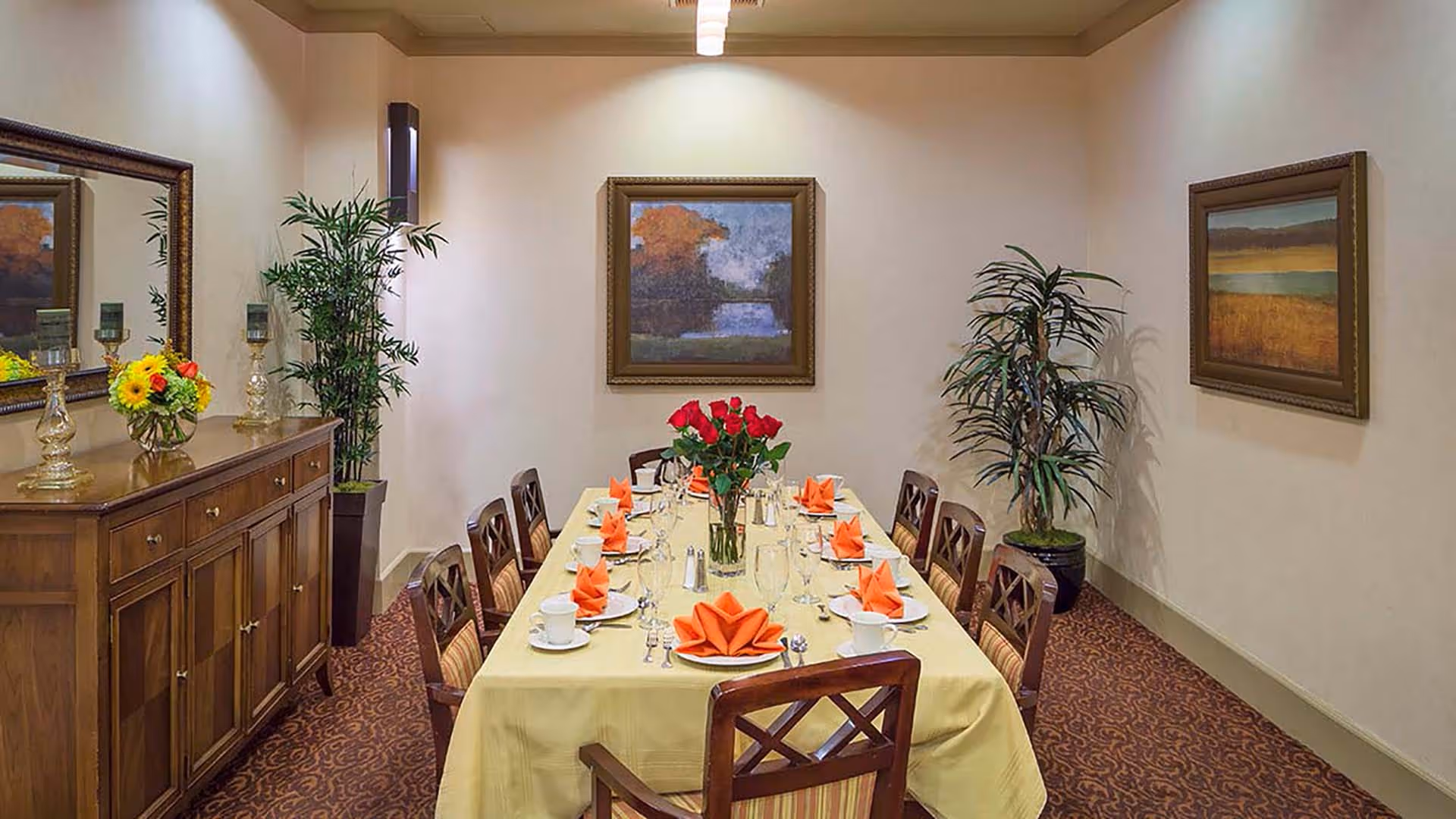 A formal dining room with a long rectangular table covered with a yellow tablecloth, set with white plates, cups, silverware, wine glasses, and orange folded napkins. A vase with red roses is placed in the center of the table. The room has patterned carpet, two framed landscape paintings on the walls, a wooden sideboard with decorative items including a flower arrangement and candle holders, and two potted plants in the corners.