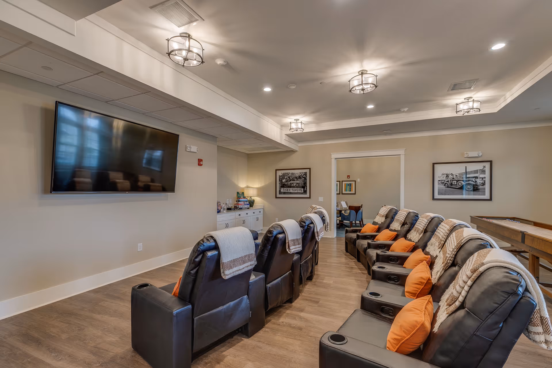 A cozy entertainment room with two rows of black leather recliners, each with an orange pillow and a beige throw blanket. A large flat-screen TV is mounted on the wall opposite the chairs. The room has wood flooring, soft beige walls, and ceiling lights with decorative fixtures. There are framed pictures on the walls and a shuffleboard table to the right side of the room.