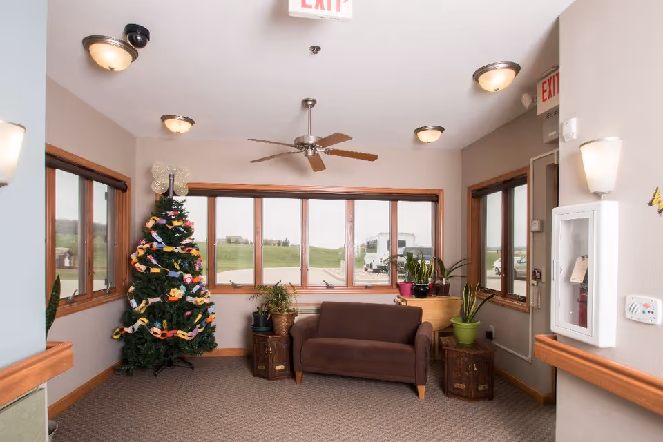 Small seating area with a brown sofa, decorated Christmas tree, potted plants, and large windows.