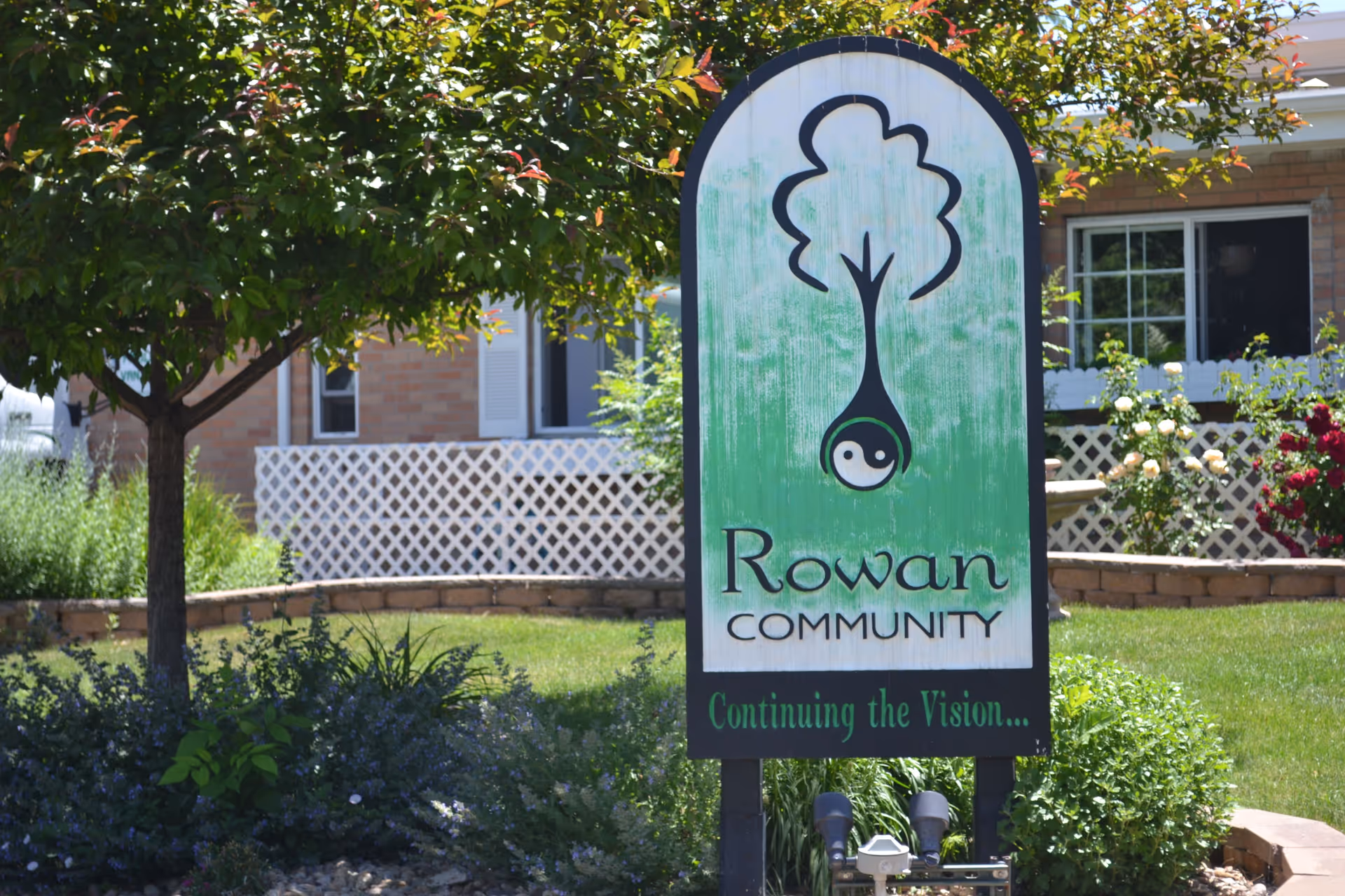 A green and white 'Rowan Community' sign stands in a landscaped front yard with a tree, flowers, and the building facade behind it.
