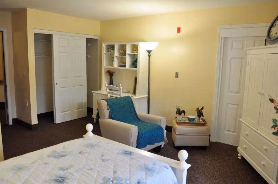 A cozy bedroom with a white bed featuring a floral quilt, a beige armchair draped with a blue blanket, a white desk with shelves holding decorative items, a floor lamp, and a white wardrobe. There are two closets with sliding doors and a small ottoman with a tray on top.