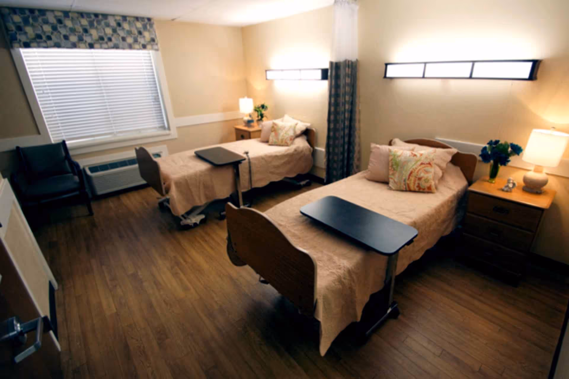 A nursing facility room with two single beds, each with beige bedspreads and decorative pillows. Each bed has an overbed table. There are two nightstands with lamps and a vase of flowers. A window with blinds and a patterned valance is on the left wall, and two modern wall lights are above the beds. The floor is wooden, and there is a dark armchair near the window.