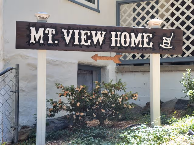 Wooden sign reading "MT. VIEW HOME" with an arrow and rocking chair motif standing in front of a building and landscaping.