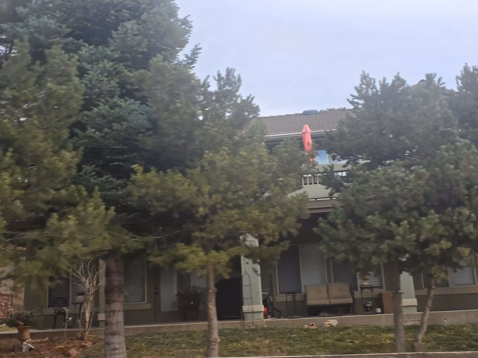 Exterior view of a residential building partially obscured by several evergreen trees. The building has a covered porch with outdoor seating and a balcony above with a red umbrella.