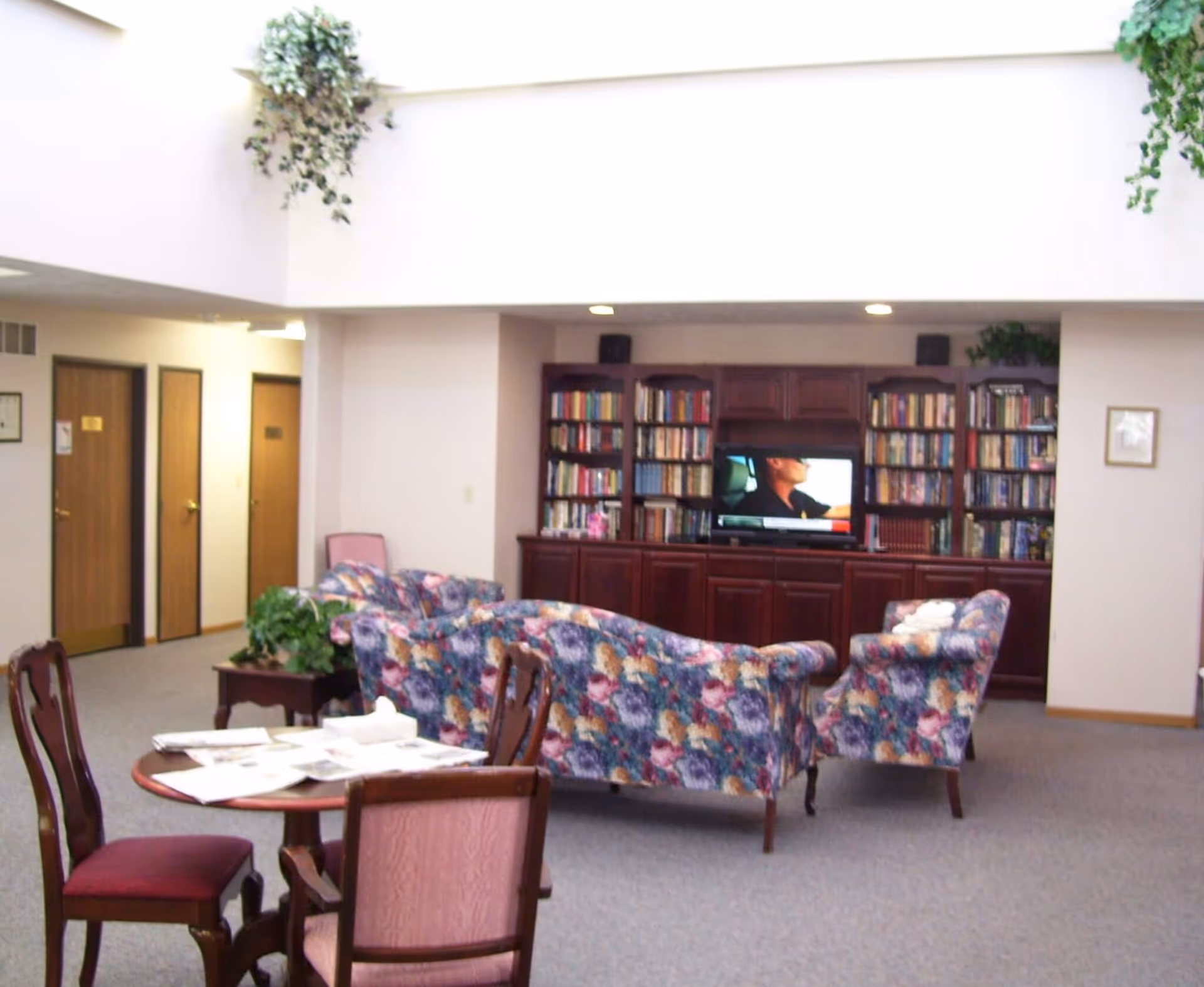 A cozy living room area with floral patterned sofas and armchairs arranged around a television set on a wooden cabinet filled with books. There is a small round table with chairs and newspapers in the foreground. The room has a high ceiling with plants hanging from the upper ledge and several wooden doors along the wall.
