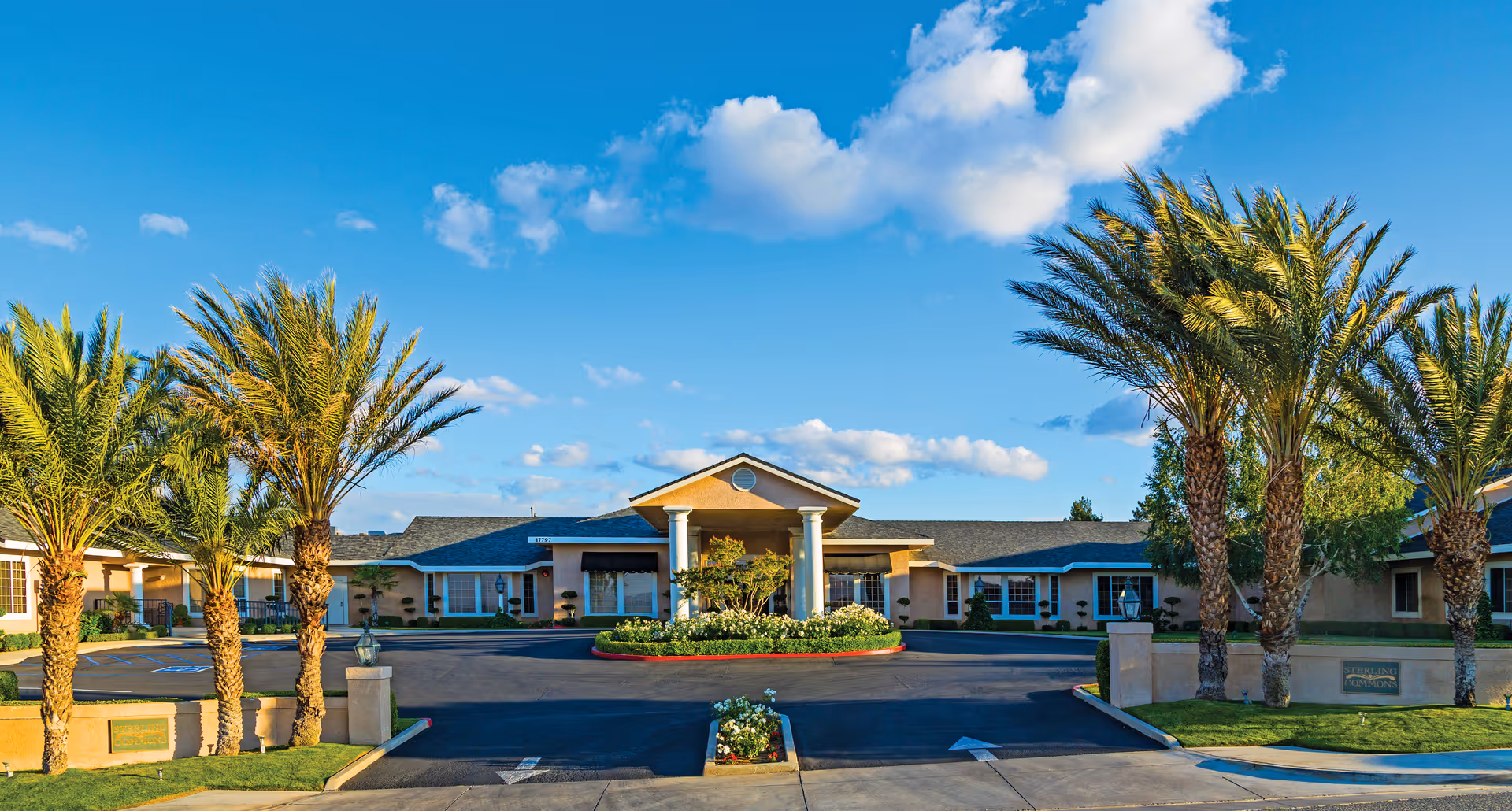 Front exterior view of Sterling Commons Memory Care Community building with a driveway, landscaped garden with flowers, and palm trees under a blue sky with some clouds.