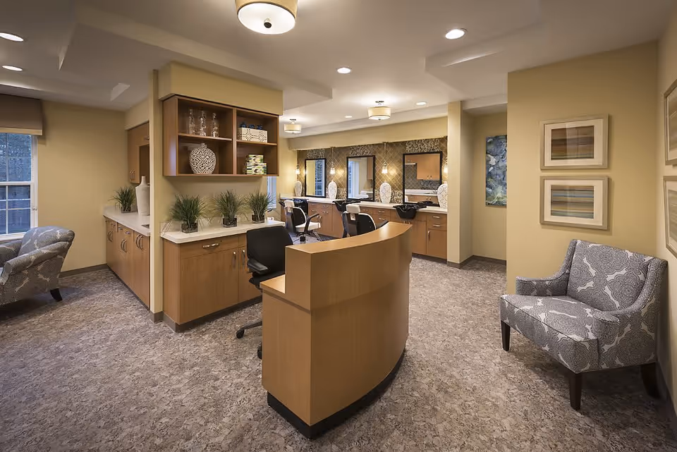 Interior view of a salon area in a senior living facility with a curved wooden reception desk, salon chairs, mirrors, and cabinetry. The room has beige walls, patterned armchairs, decorative plants, and framed artwork on the walls.