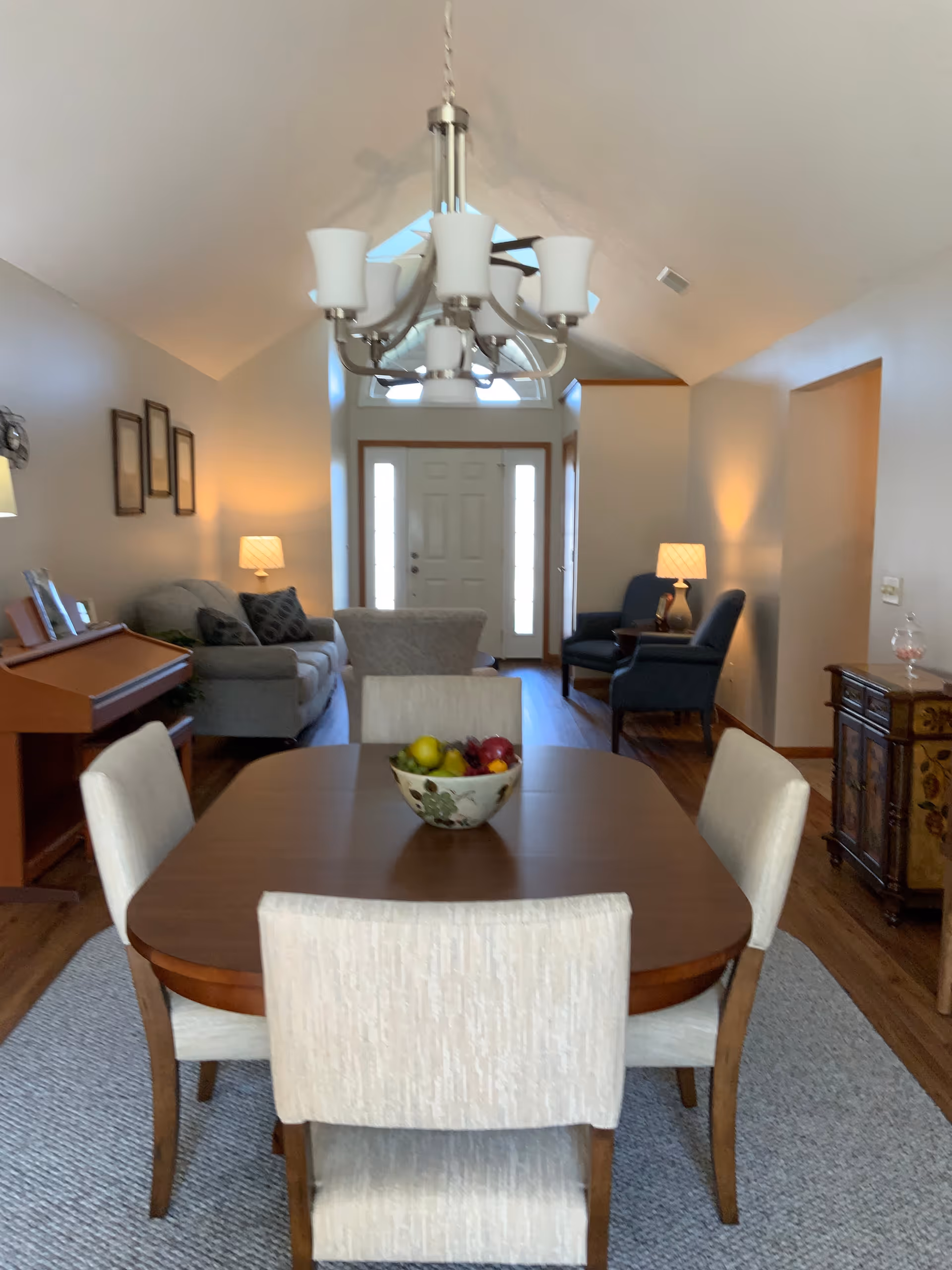 Interior view of a living and dining area in a retirement community. The foreground features a wooden dining table with four upholstered chairs and a decorative bowl of fruit on top. In the background, there is a living room with a sofa, armchairs, side tables with lamps, and a piano against the left wall. The space has wooden flooring, a high vaulted ceiling with a chandelier, and a front door with sidelights and a transom window letting in natural light.