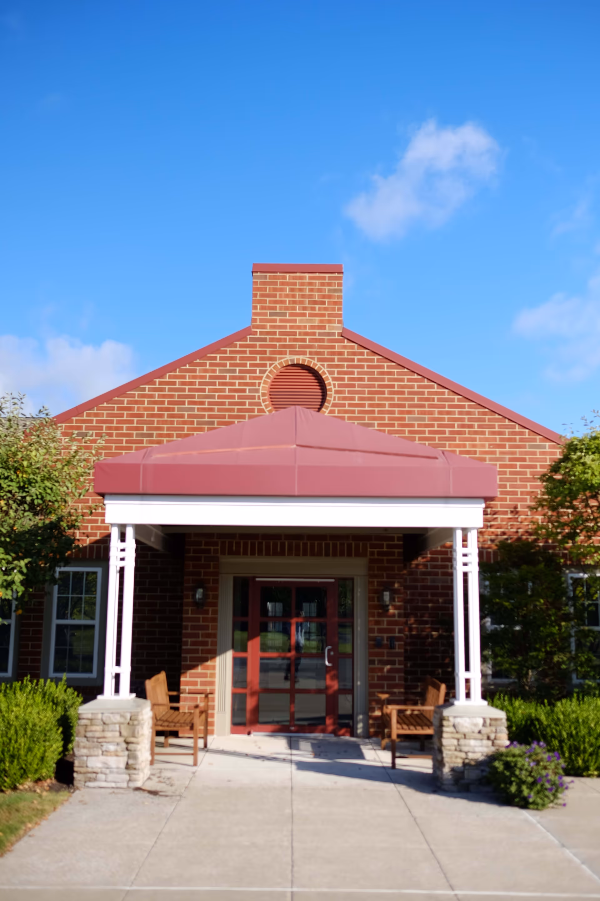 Front entrance of a brick building with a red peaked roof over the doorway, two wooden benches on either side of the entrance, and green bushes and trees surrounding the walkway leading up to the door under a clear blue sky.