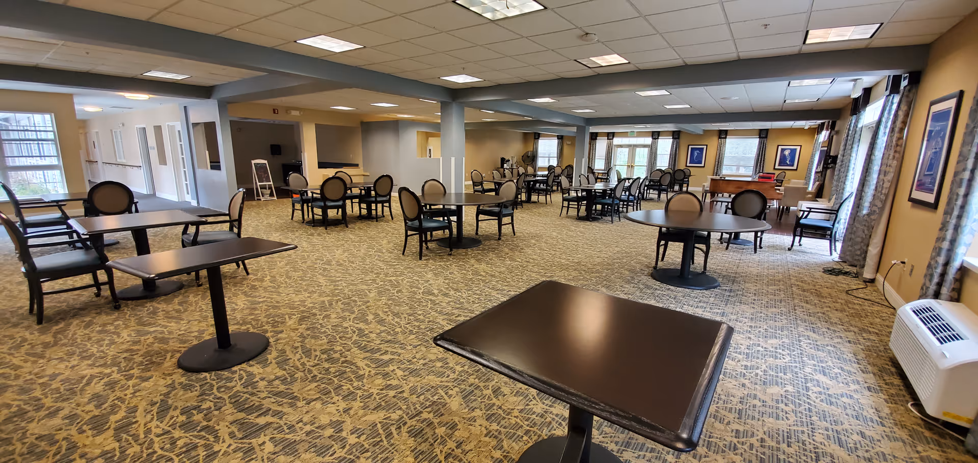 Spacious senior living dining/activity room with multiple round and rectangular tables and chairs on patterned carpet under a drop ceiling.