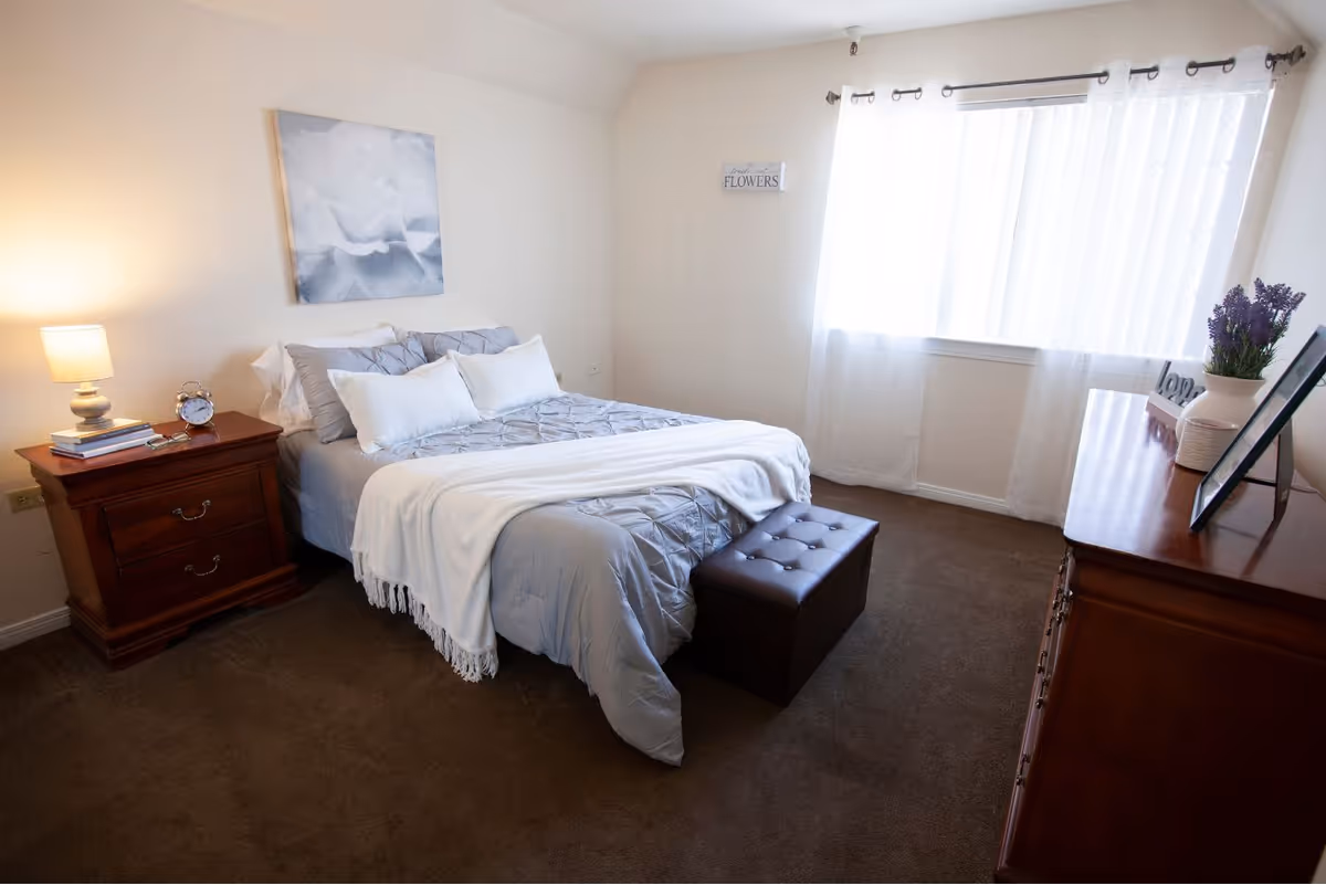 Neatly furnished bedroom with a made bed, nightstand with a lamp, dresser, and a window with sheer curtains.