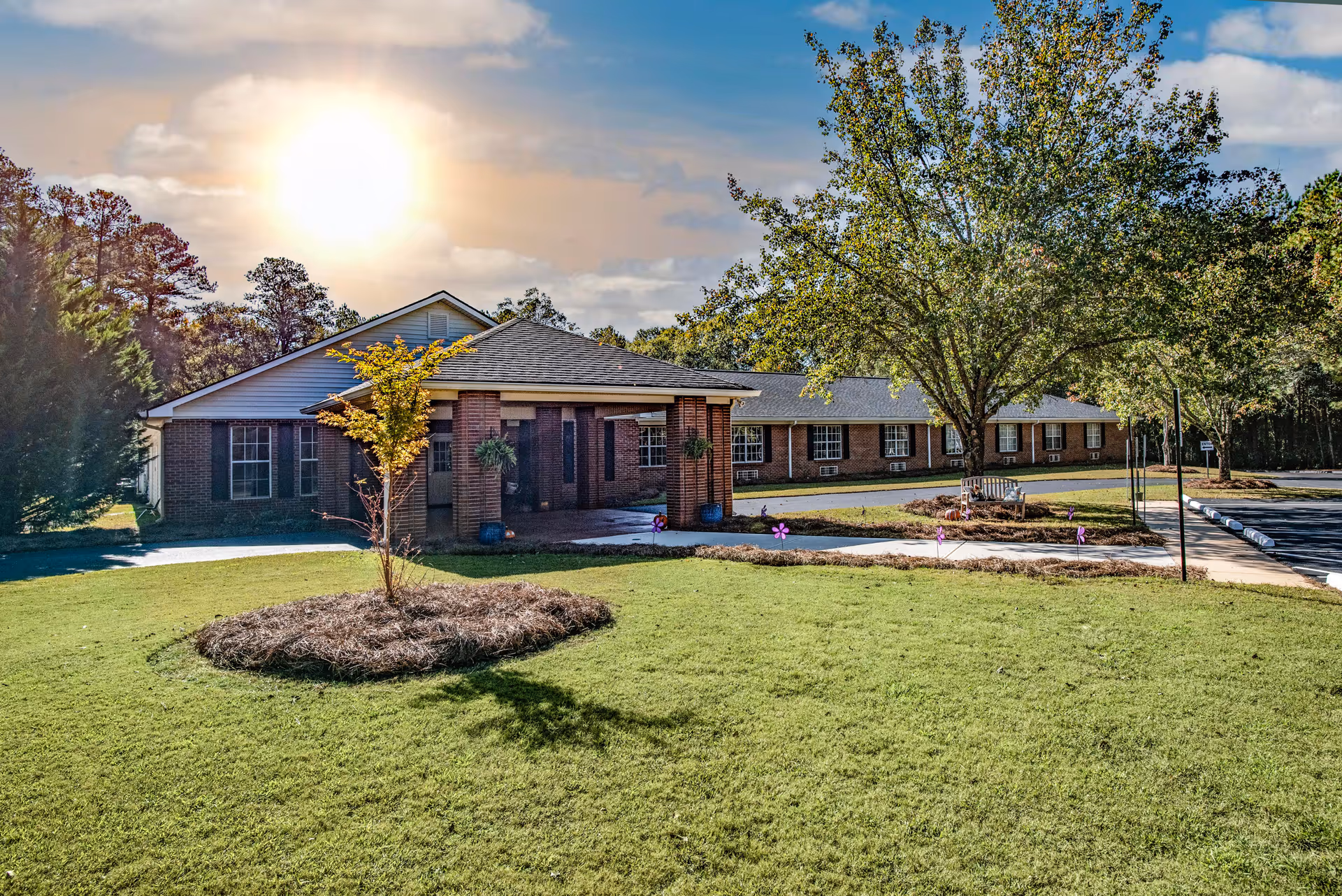 Exterior view of a single-story brick building with a covered entrance, surrounded by green grass, trees, and a clear sky with the sun shining brightly.