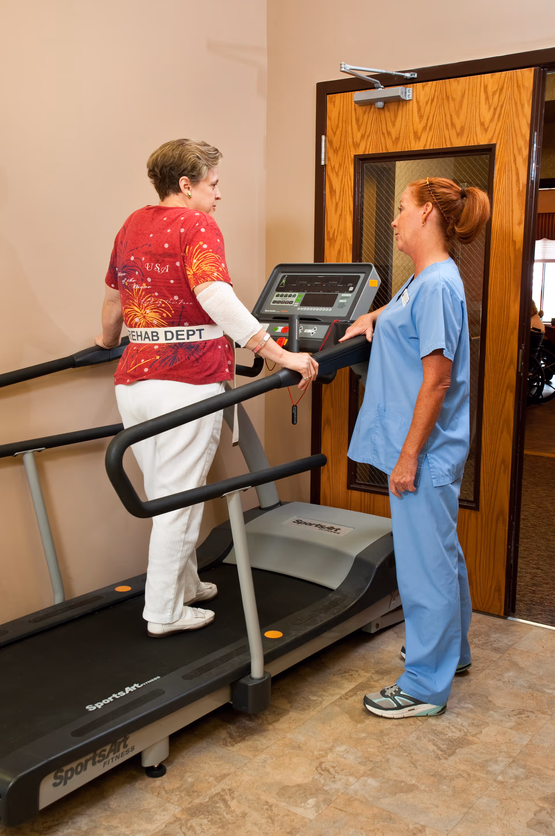 A woman wearing a red shirt with a 'REHAB DEPT' belt is walking on a treadmill while a healthcare professional in blue scrubs stands beside her, providing assistance. They are in an indoor room with a wooden door in the background.