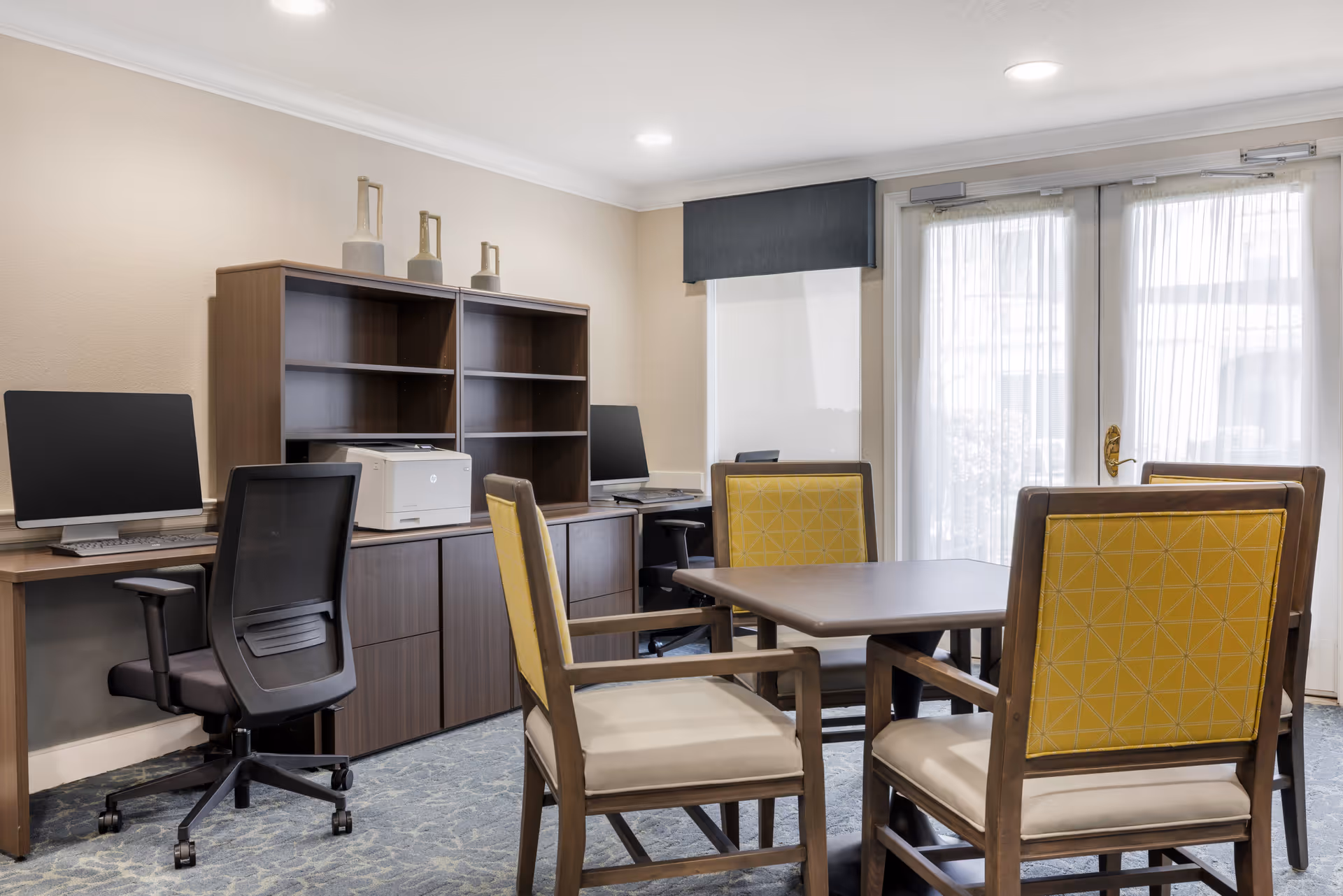 A well-lit office area with two computer workstations on a wooden desk, a printer, and a wooden shelving unit. In the foreground, there is a square table surrounded by four wooden chairs with yellow patterned backs and beige seats. The room has carpeted flooring, light-colored walls, and large glass doors with sheer curtains allowing natural light to enter.