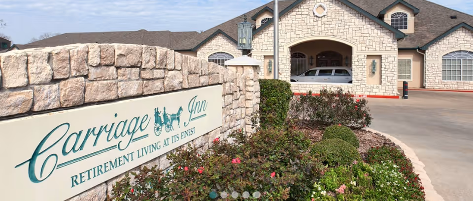 Stone sign for 'Carriage Inn Retirement Living at Its Finest' by landscaped shrubs with the facility's stone-front building and driveway in the background.