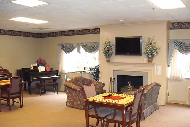 A cozy common area in Briarwood Health Center featuring a fireplace with a TV mounted above it, two floral-patterned armchairs, a table with a checkers board, a piano in the corner, and windows with decorative curtains letting in natural light.