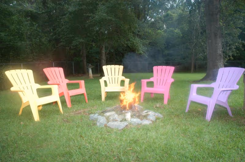 A circle of colorful Adirondack chairs in yellow, red, pink, and purple arranged around a small campfire on a grassy lawn with trees and a fence in the background.