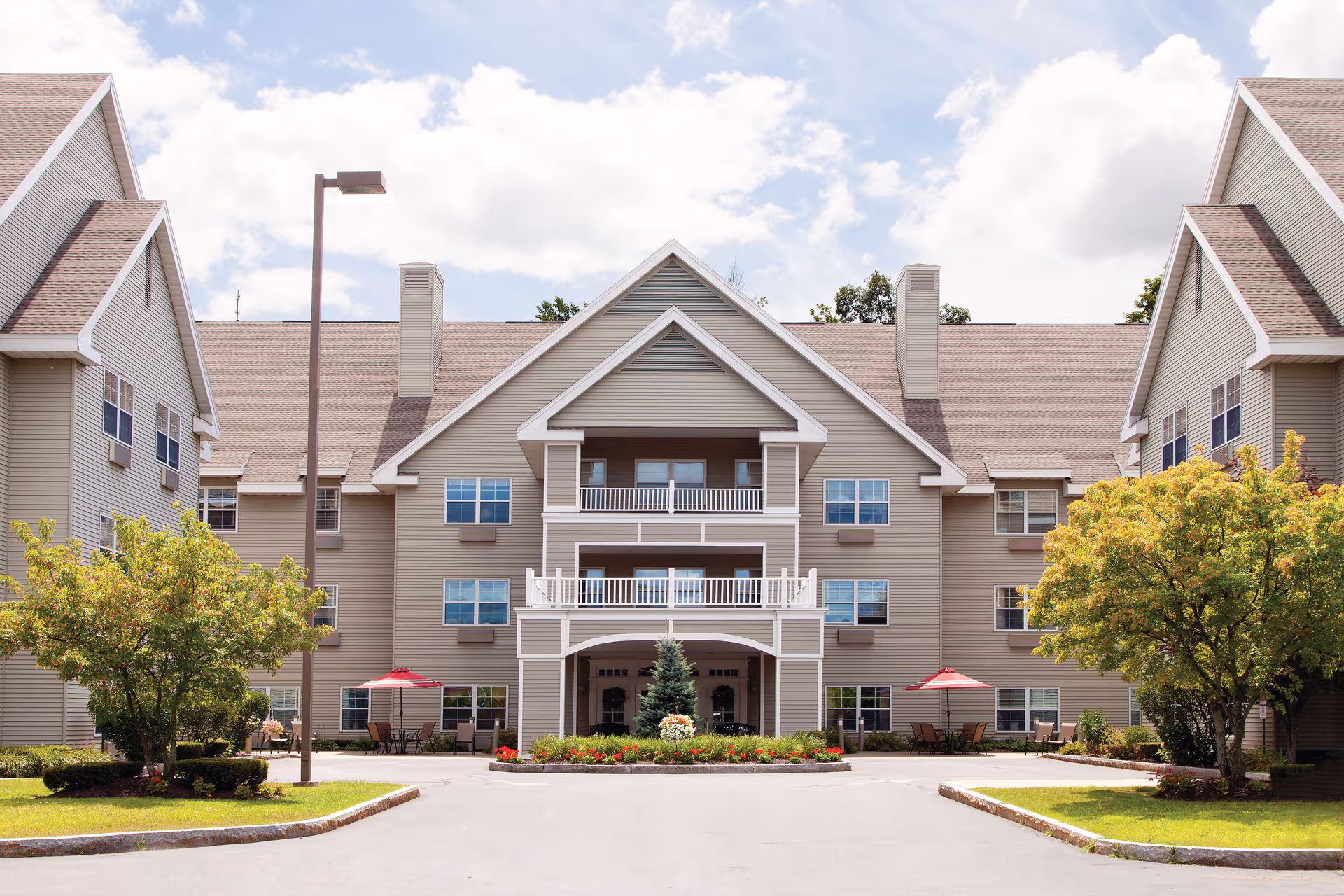 Front exterior view of a large, multi-story senior living facility building with beige siding, multiple windows, and a central entrance with balconies above. There are landscaped areas with trees and flowers, outdoor seating with red umbrellas, and a clear sky with some clouds.