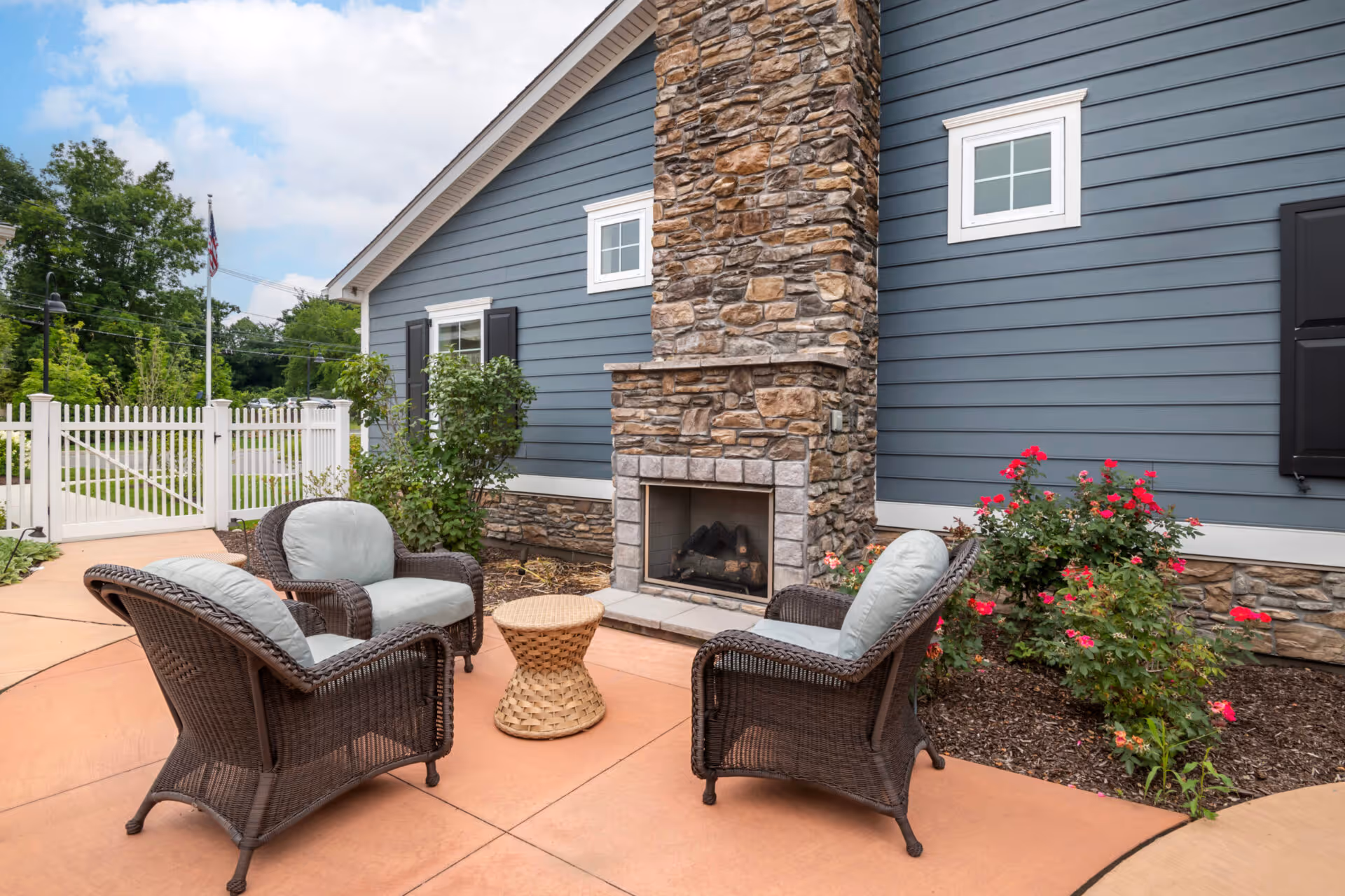 Outdoor patio area with four cushioned wicker chairs arranged around a small woven table in front of a stone fireplace attached to a blue building with white trim. There are flowering bushes and a white picket fence in the background under a partly cloudy sky.