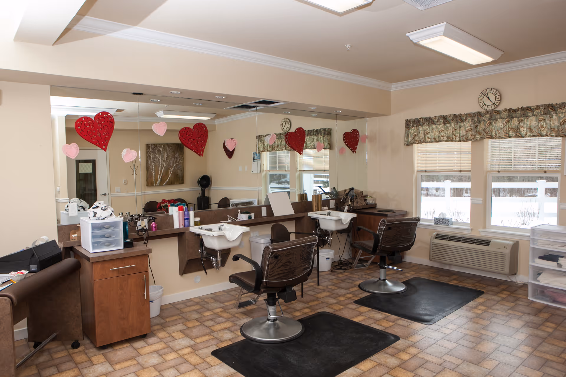 Interior of a hair salon area with two styling chairs in front of sinks and mirrors. The room has beige walls, a patterned floor, and windows with floral valances. Red and pink heart decorations hang on the mirrors.