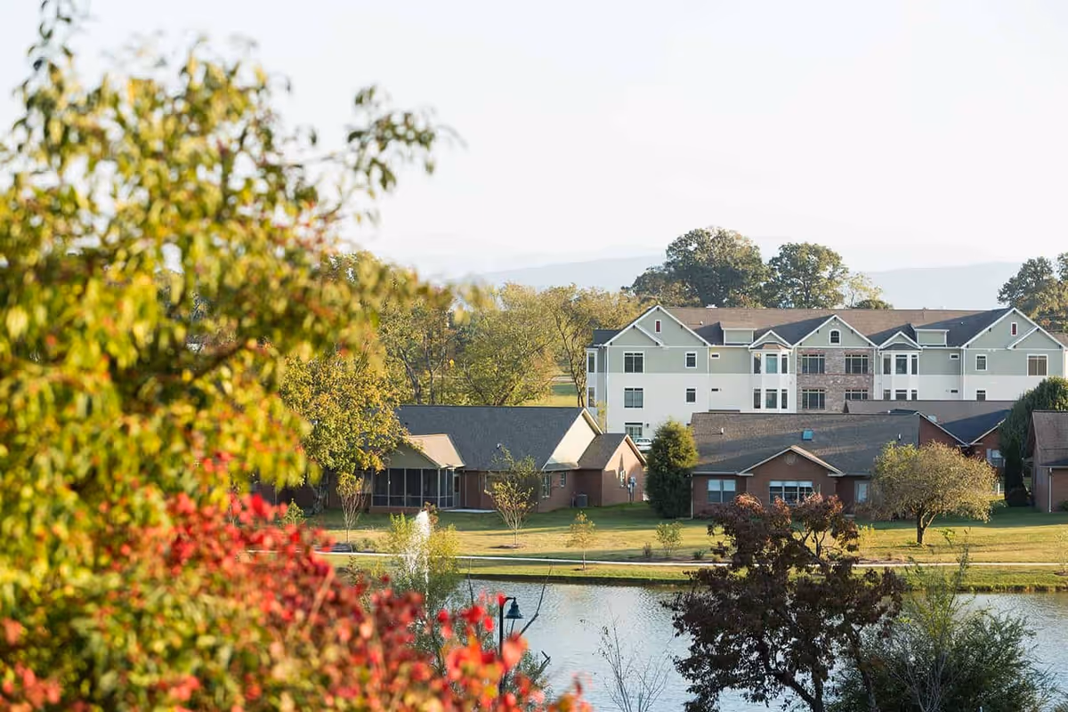 View of a senior living facility with multiple buildings surrounded by trees and greenery, with a pond in the foreground and a fountain in the water.