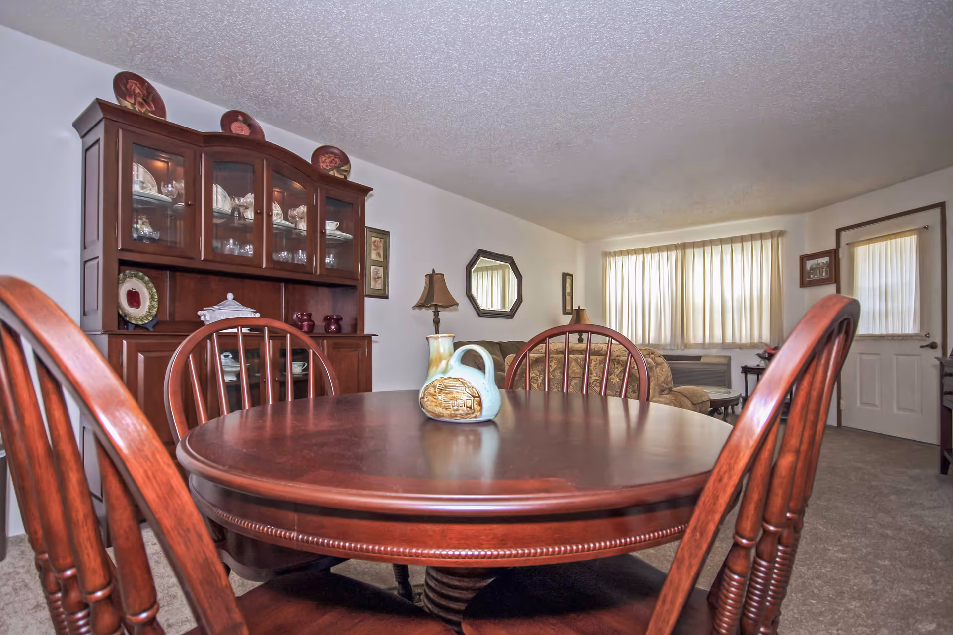 Interior view of a senior living facility showing a wooden dining table with four chairs and a decorative ceramic pitcher on top. In the background, there is a wooden china cabinet with glass doors displaying dishes and glassware, a sofa, two table lamps, a mirror on the wall, a window with curtains, and a door.