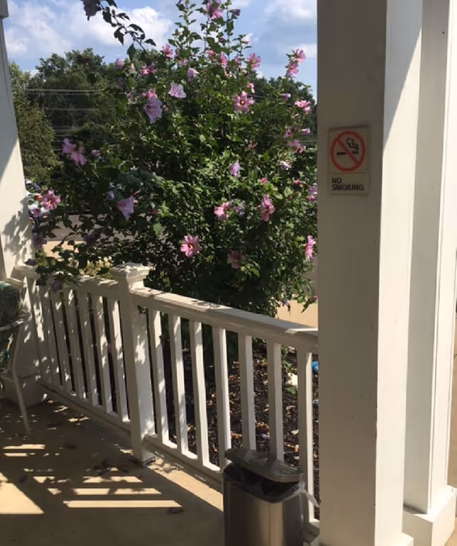 View of a porch area with white railing and a blooming bush with purple flowers. A no smoking sign is posted on a white pillar, and a trash can is placed near the pillar. The background shows a parking lot and trees under a partly cloudy sky.