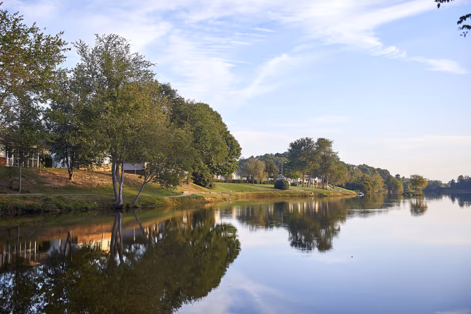 Calm lakeside view showing trees, grassy shoreline and houses reflected in still water under a partly cloudy sky.
