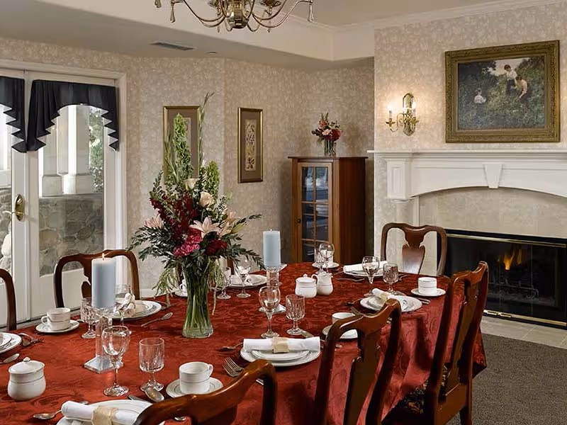 A formal dining room with a long table covered in a red tablecloth set with white plates, cups, glasses, silverware, and candles. A large floral arrangement is in the center of the table. The room features wooden chairs, a fireplace with a painting above it, wall sconces, and a glass-front cabinet. French doors with black valances lead outside.