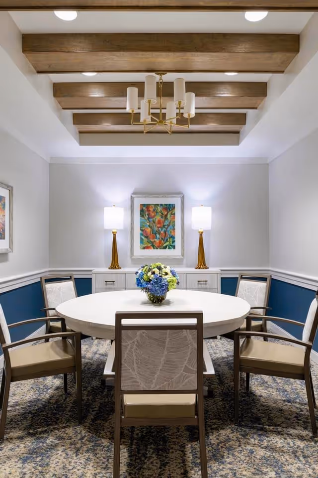 A well-lit dining room with a round white table surrounded by six cushioned chairs. The room features a patterned carpet, two table lamps with gold bases on a sideboard, a colorful floral painting on the wall, and wooden ceiling beams with a modern chandelier hanging in the center.