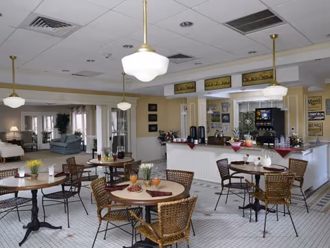 A dining area in a senior living facility featuring round tables with wicker chairs, set with glasses and small plants. The room has a tiled floor, pendant lights hanging from the ceiling, and a counter area with coffee dispensers and a beverage machine. In the background, there is a lounge area with sofas and large windows letting in natural light.