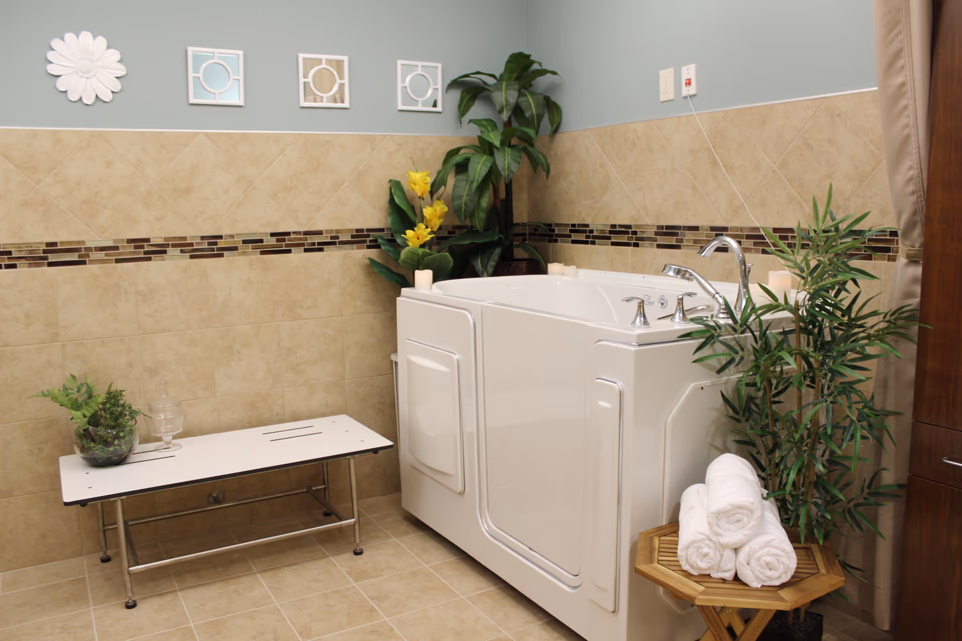 A clean and modern bathroom featuring a white walk-in bathtub with chrome faucets, surrounded by beige tiled walls with a decorative mosaic border. There are green plants placed around the tub, a small wooden table holding rolled white towels, and a white bench with a glass and a small plant on it. The wall above the tiles is painted light blue and decorated with three square mirrors and a white flower-shaped wall ornament.