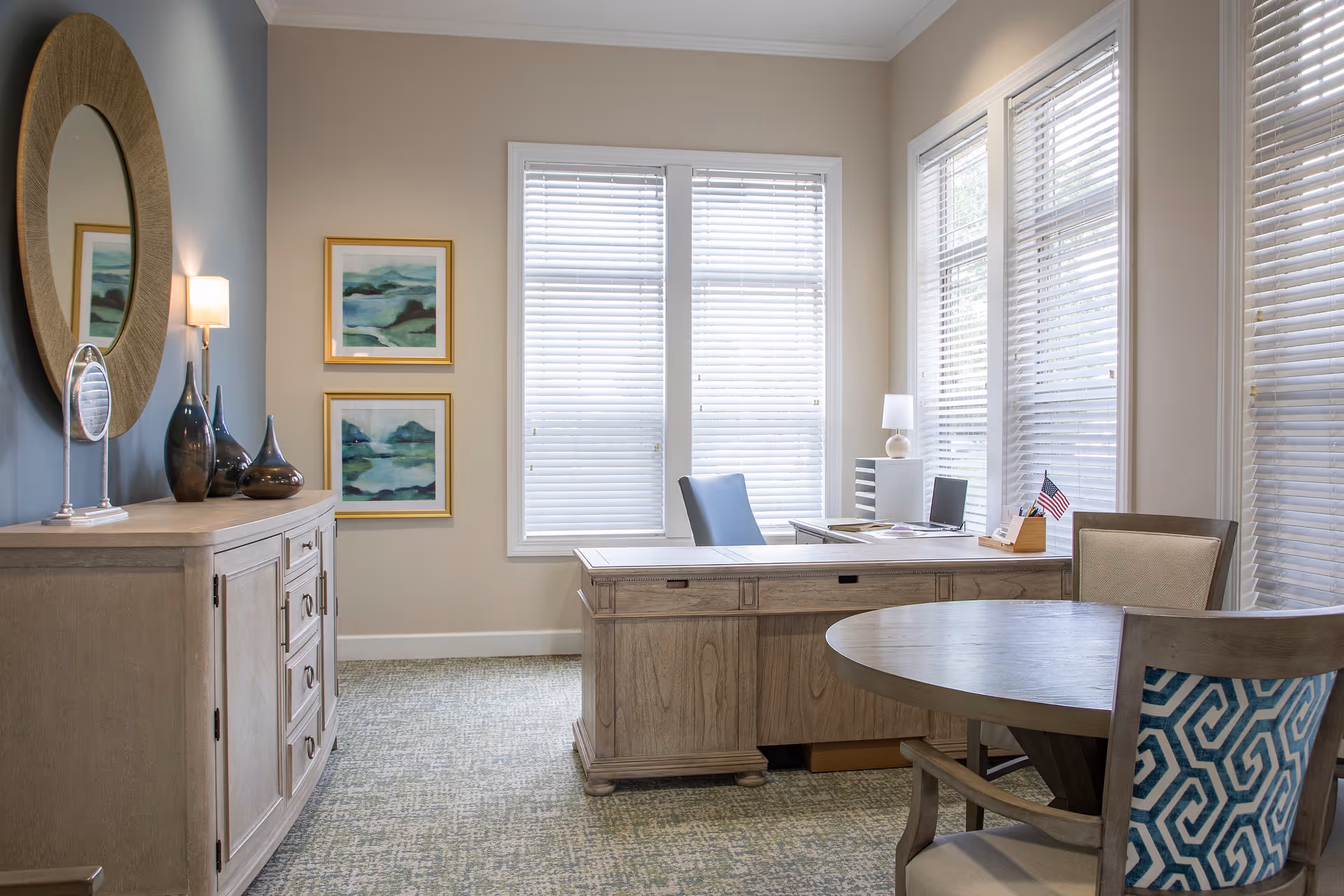 A bright office space with large windows covered by white blinds. The room features a wooden desk with a blue office chair, a round wooden table with two chairs, and a wooden sideboard with decorative vases and a round mirror above it. Two framed abstract paintings hang on the wall.