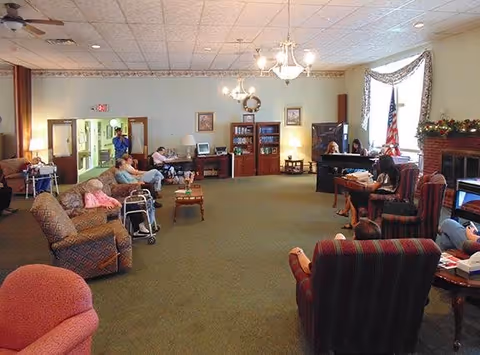 A spacious living room in a senior living facility with several elderly residents seated in armchairs and using walkers. The room features carpeted floors, a ceiling fan, a chandelier, and large windows with curtains. There is a bookshelf, a television, and an American flag near a fireplace decorated with garland. Staff members are present, assisting or interacting with residents.