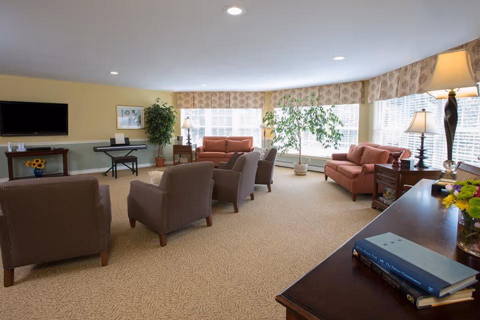 A spacious senior living common area with multiple armchairs and sofas arranged around the room. There are large windows with patterned valances letting in natural light, potted plants, table lamps, a flat-screen TV mounted on the wall, and a keyboard on a stand. A wooden table with books and a vase of flowers is visible in the foreground.