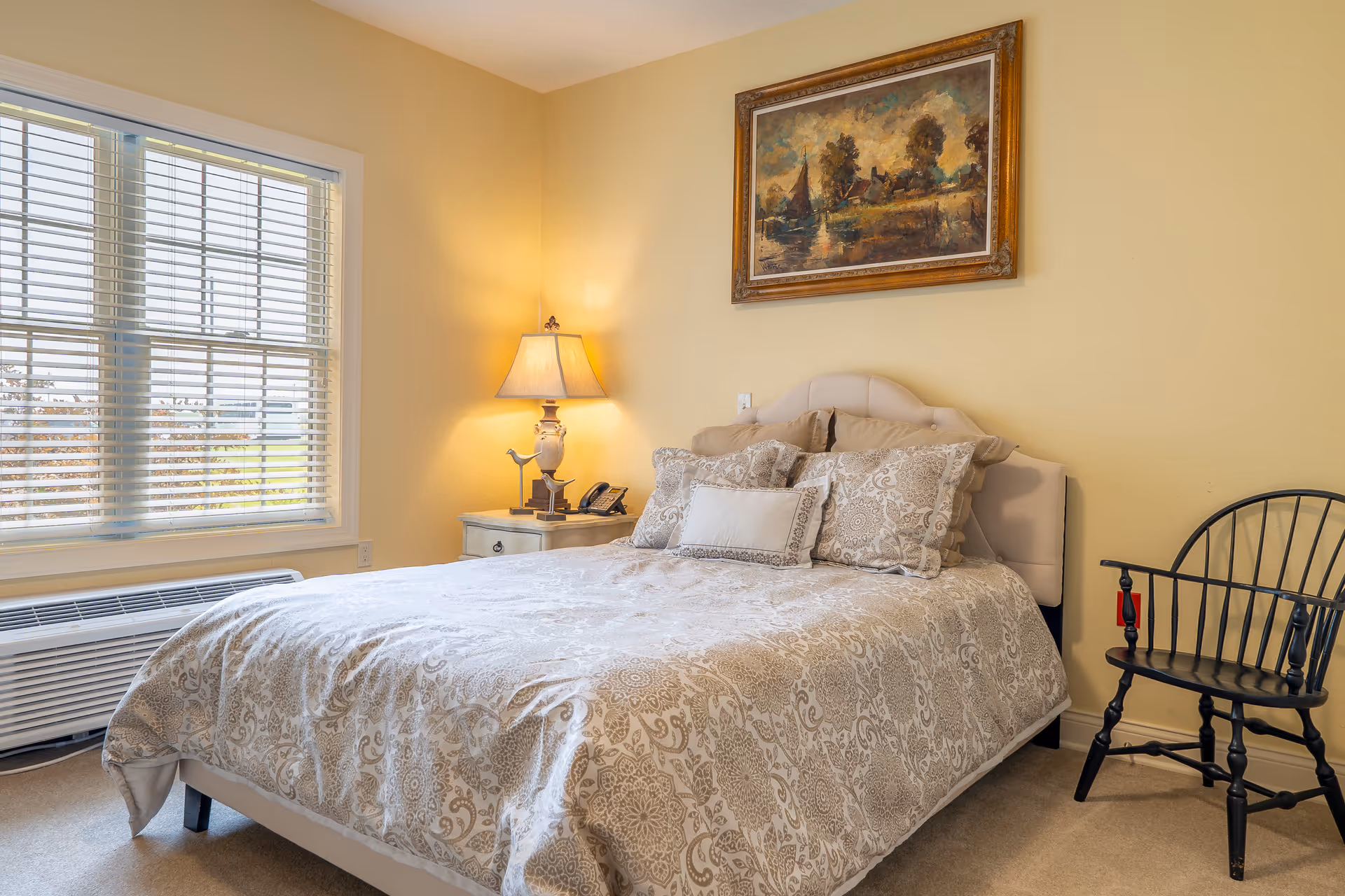 A cozy bedroom with a large bed covered in a patterned beige and white comforter and multiple pillows. There is a nightstand with a lamp and a telephone next to the bed. A framed painting depicting a landscape with trees and a boat hangs on the yellow wall above the bed. A black wooden chair is placed against the wall to the right of the bed. A large window with white blinds allows natural light into the room.