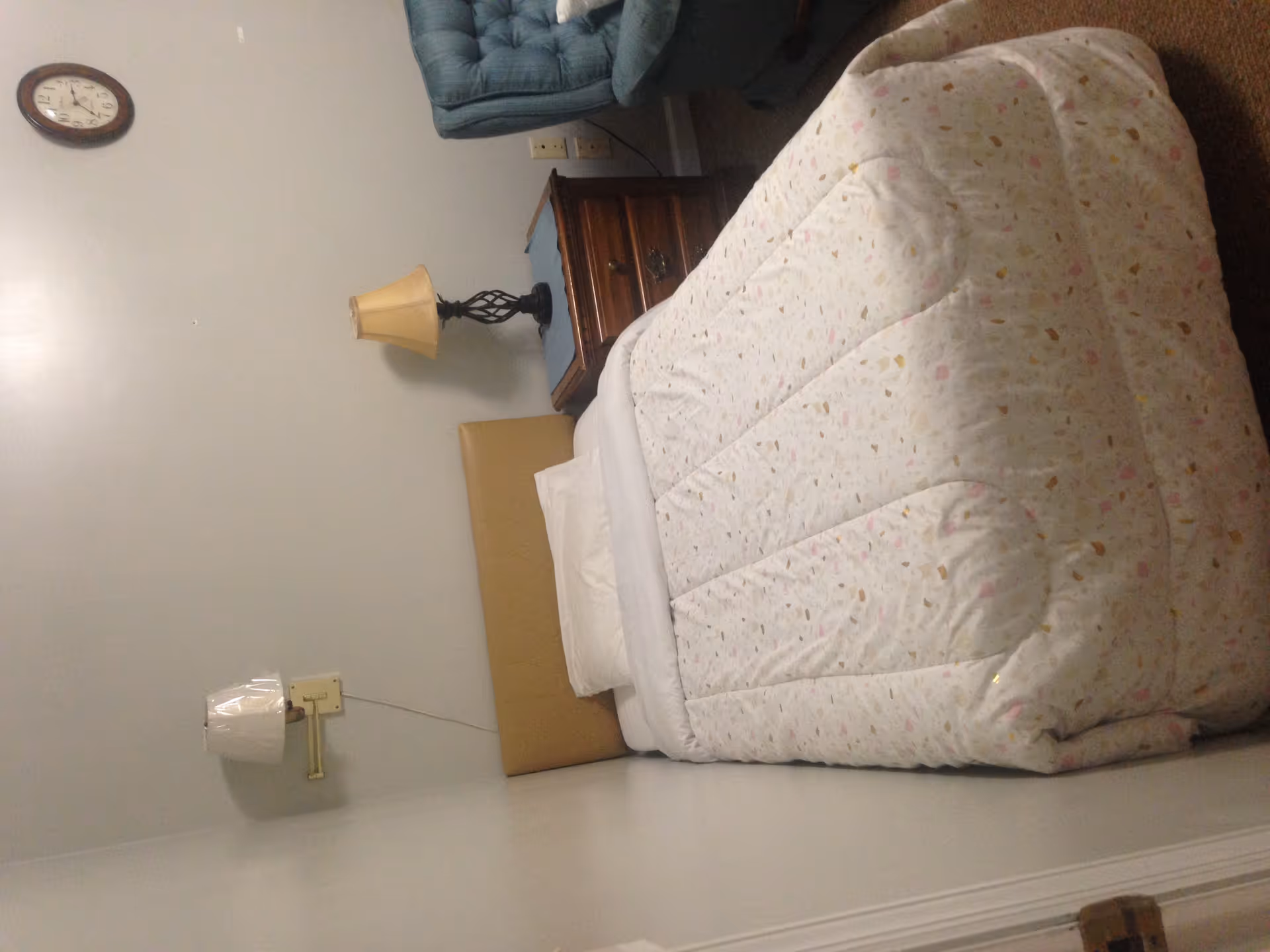 A small bedroom with a single bed covered with a white patterned comforter and a white pillow. There is a wooden nightstand with a lamp on it next to the bed, a wall-mounted lamp above the bed, a blue upholstered chair to the right, and a round wall clock above the nightstand on a plain light-colored wall.