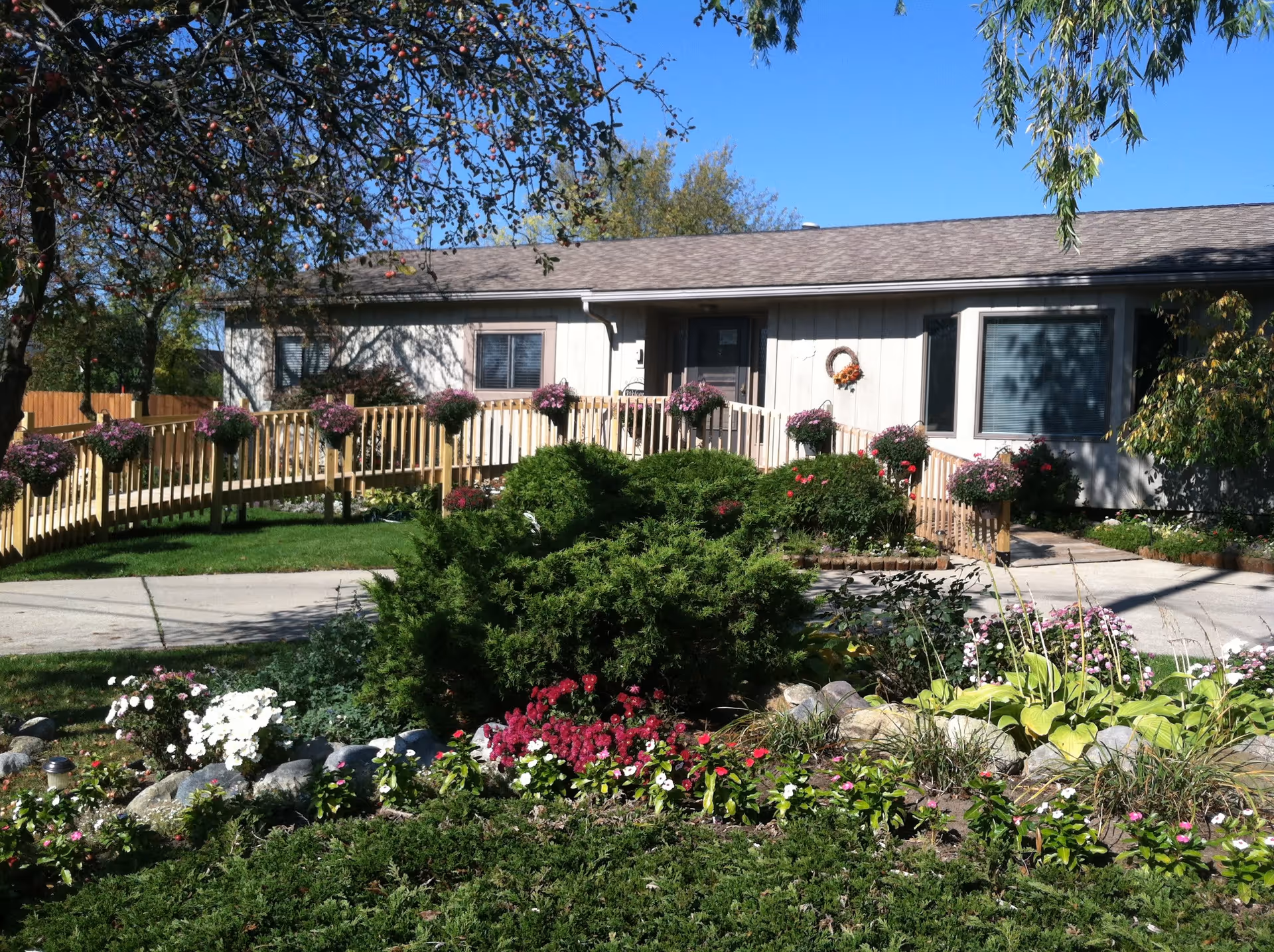 Single-story building with a sloped roof, surrounded by a well-maintained garden featuring various flowers and shrubs. A wooden ramp with flower pots on the railing leads to the entrance door. Trees and a clear blue sky are visible in the background.