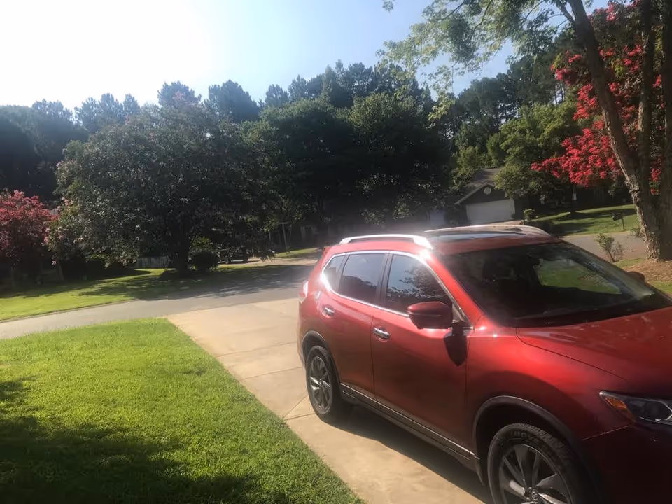 Red SUV parked in a driveway beside a green lawn with trees and houses on a sunny suburban street.