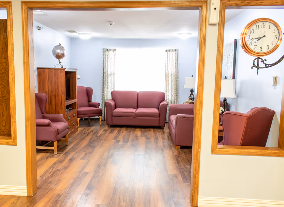 A cozy living room with wooden flooring, featuring a maroon loveseat, two matching armchairs, a wooden entertainment cabinet with a globe on top, and a wall clock. The room is softly lit with ceiling lights and natural light coming through a window with patterned curtains.