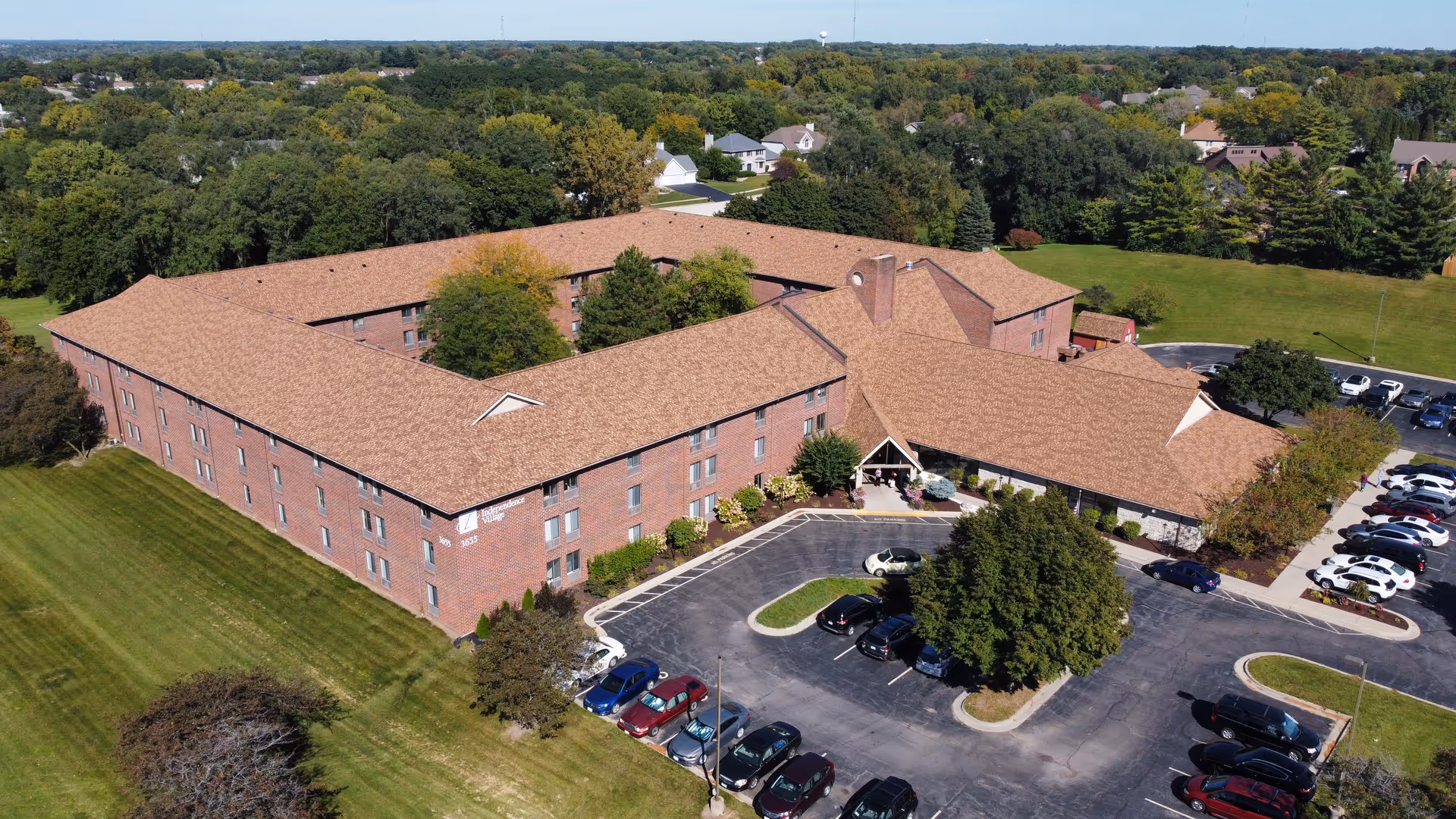 Aerial view of a large, three-story brick senior living facility named Independence Village, surrounded by green lawns, trees, and a parking lot with several cars. The building has a tan shingled roof and is situated in a suburban area with other houses and trees in the background.