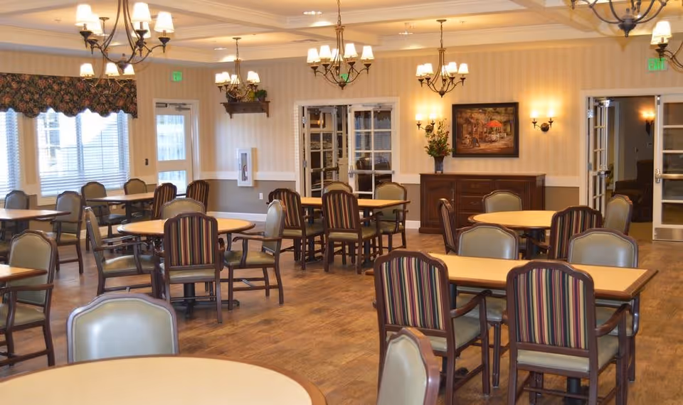 A spacious dining room in Colonial Gardens Assisted Living & Memory Care featuring multiple round and rectangular tables with cushioned chairs. The room has wooden flooring, soft yellow walls with white trim, several chandeliers and wall sconces providing warm lighting, and large windows with floral valances allowing natural light to enter. A sideboard with a flower arrangement and a framed painting is visible on one wall.
