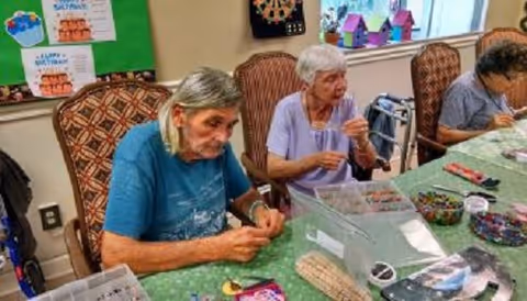 Three elderly women sitting at a table engaged in a bead crafting activity in a well-lit room with patterned chairs and a bulletin board in the background.