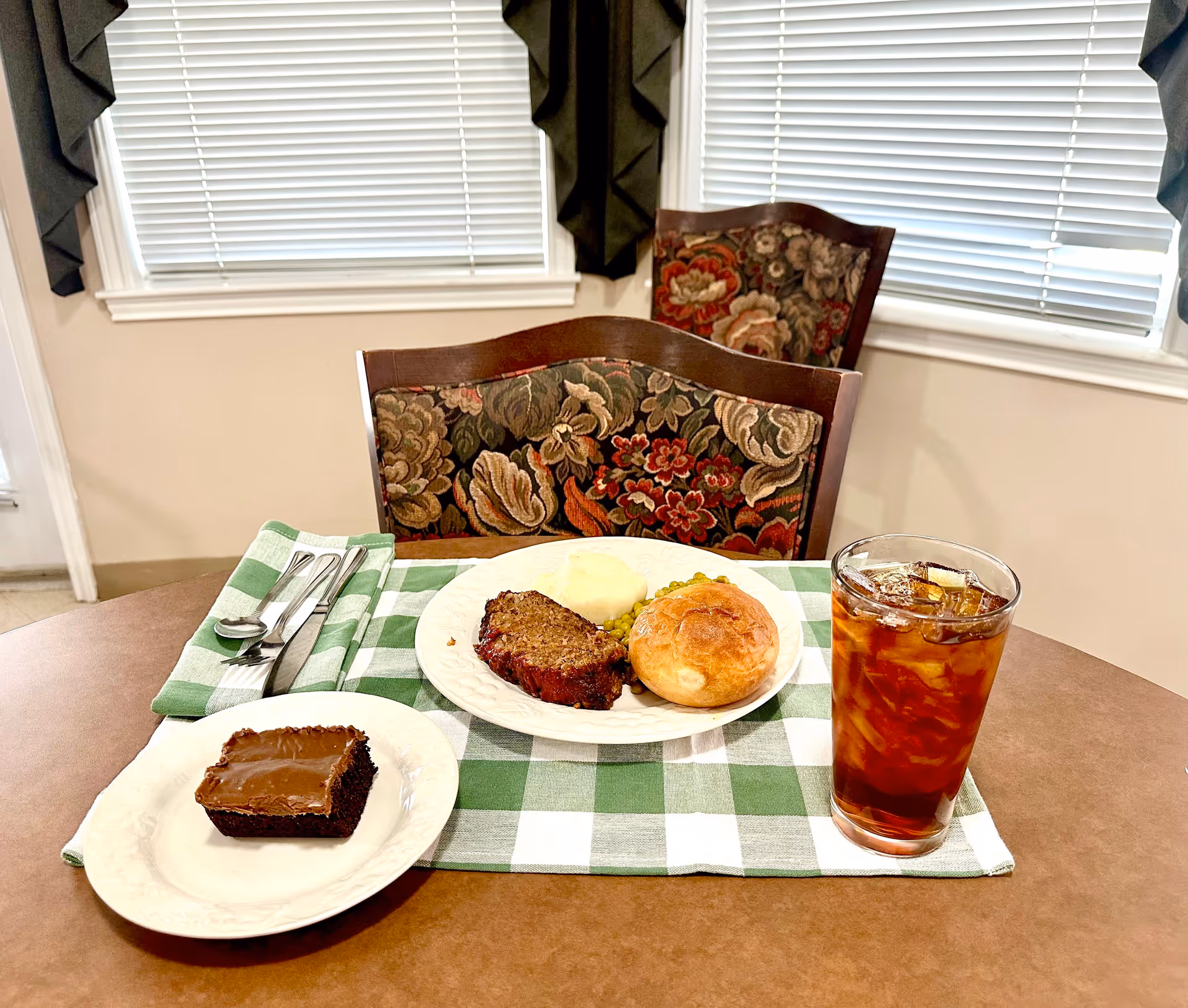 A dining table set with a green and white checkered placemat, a plate of meatloaf, mashed potatoes, peas, and a bread roll, a glass of iced tea, a small plate with a chocolate frosted brownie, and silverware wrapped in a green and white napkin. Two floral upholstered chairs and windows with blinds and dark curtains are in the background.