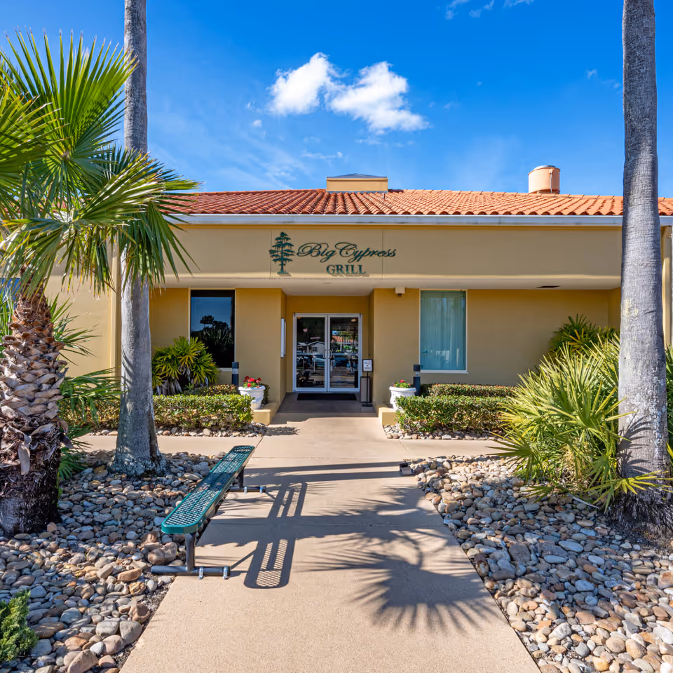 Exterior view of the Big Cypress Grill at Cypress Lakes Village, showing a beige building with a red tile roof, palm trees, shrubs, and a green bench along a walkway leading to the entrance under a blue sky with a few clouds.