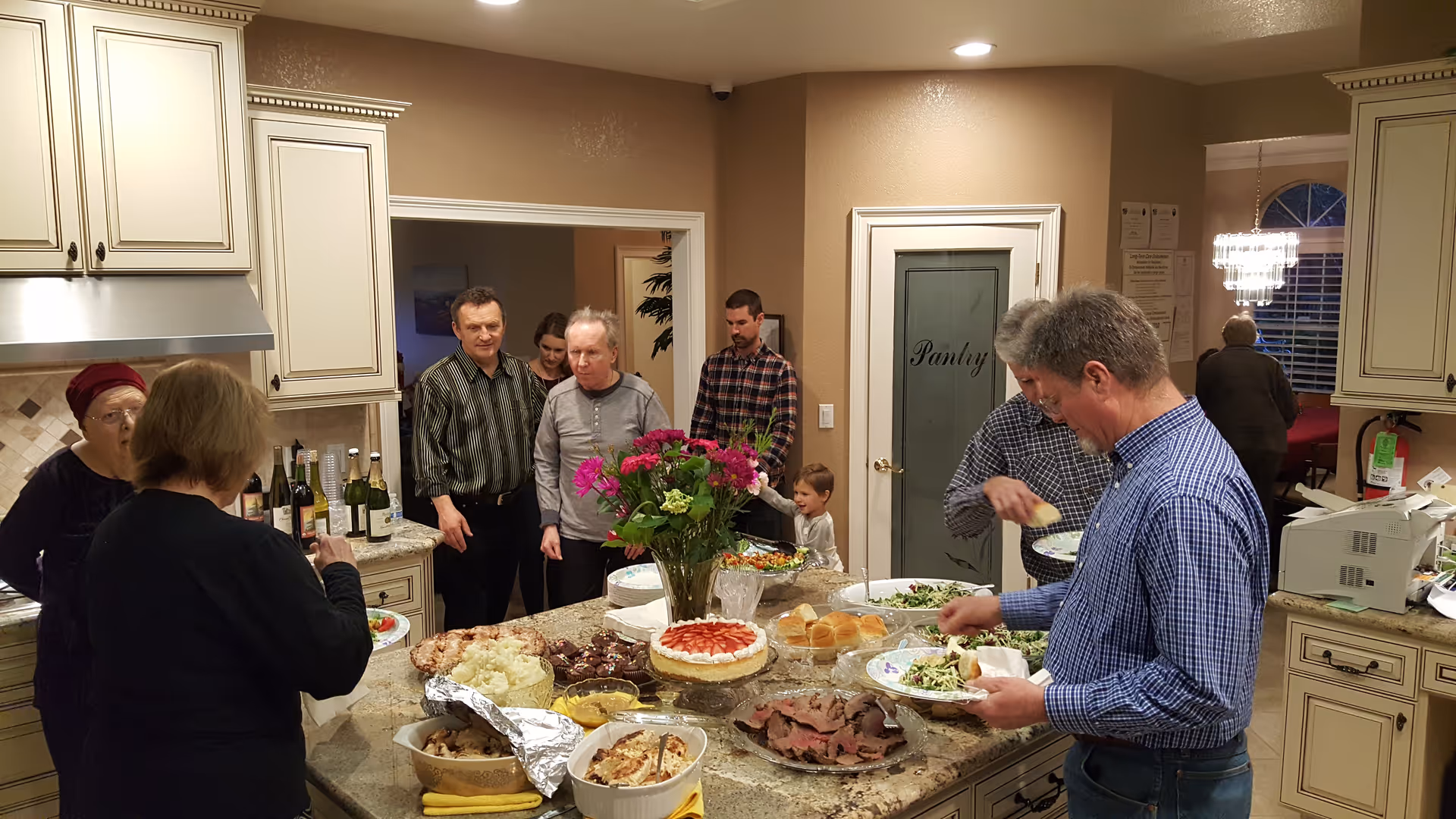 A group of people gathered in a kitchen around a large island countertop filled with various dishes including salad, roast beef, rolls, and a cake. The kitchen has cream-colored cabinets, a pantry door, and a vase with flowers on the island. Some people are serving themselves food while others are standing and talking.