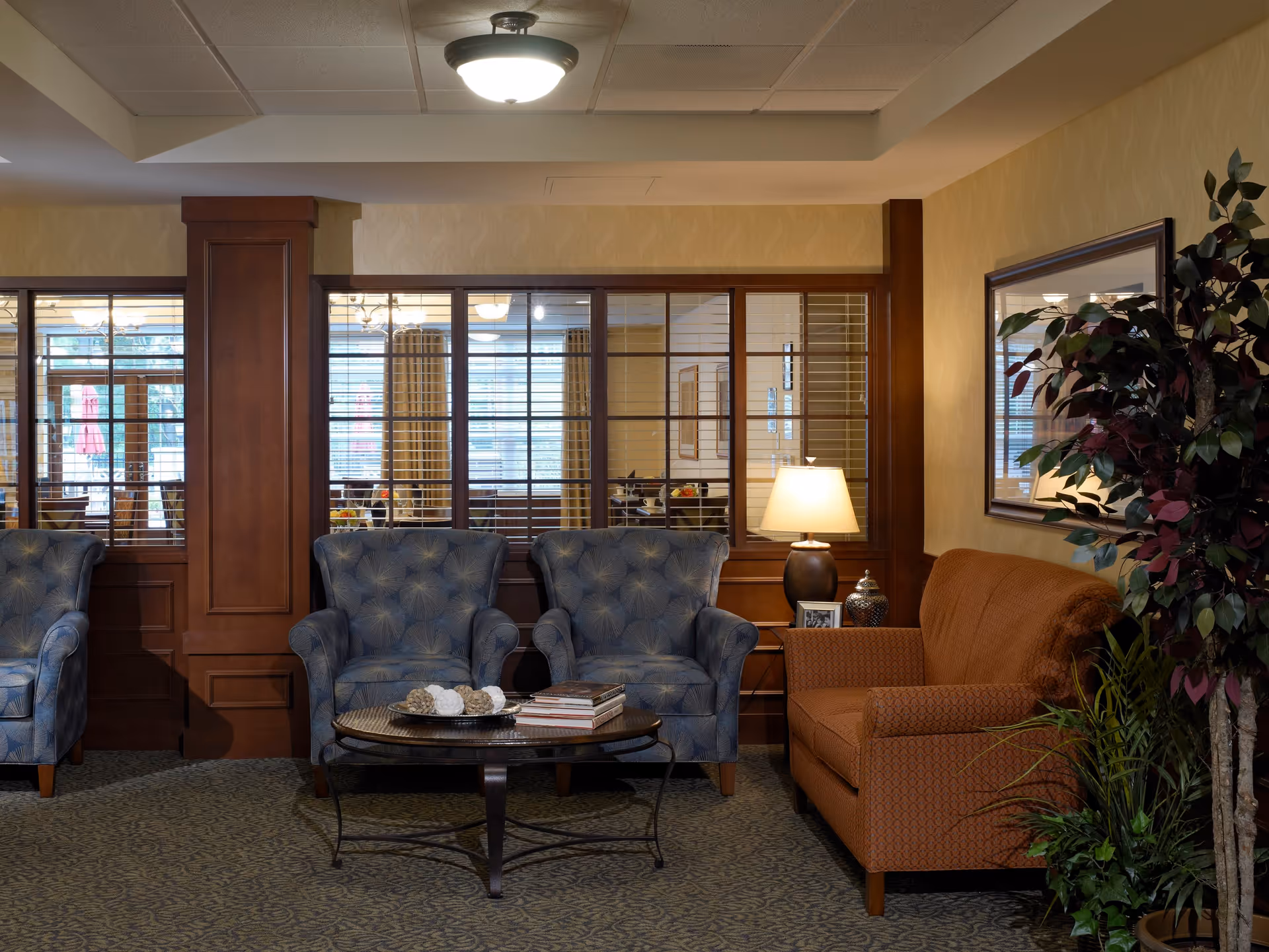 A cozy sitting area in a senior living facility with two blue upholstered armchairs and one orange sofa arranged around a round coffee table. The table holds decorative items and books. A table lamp is on a side table next to the sofa, and a large potted plant is nearby. Behind the seating area is a wooden partition with glass windows, showing a dining area with tables and chairs.