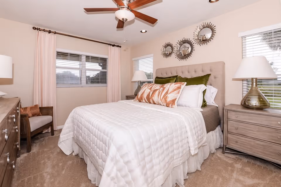 A cozy bedroom with a neatly made bed featuring white bedding and decorative pillows in green and patterned colors. The room has beige walls, a ceiling fan with wooden blades, two windows with blinds and light pink curtains, a wooden dresser on the left, a wooden nightstand with a lamp on the right, and three decorative round mirrors on the wall above the bed.