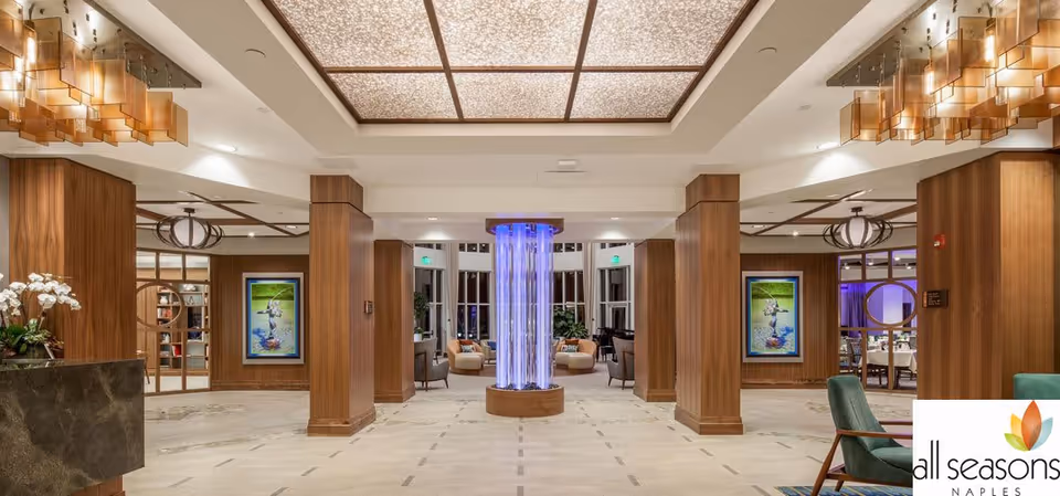 Spacious, well-lit senior living lobby with a central illuminated column, wood-paneled columns, seating areas and decorative ceiling lights.