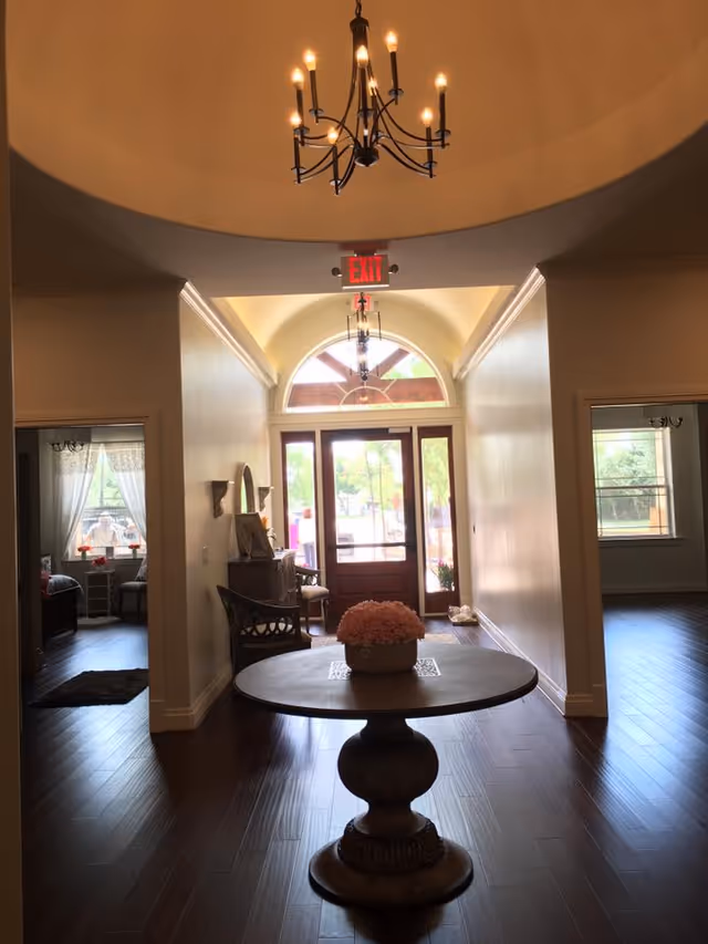 Interior view of a hallway in a senior living facility with a round wooden table holding a flower arrangement in the center. The hallway leads to a glass door with an arched window above it, letting in natural light. There are two rooms visible on either side of the hallway, each with windows and curtains. A chandelier hangs from the ceiling, and an exit sign is above the door.
